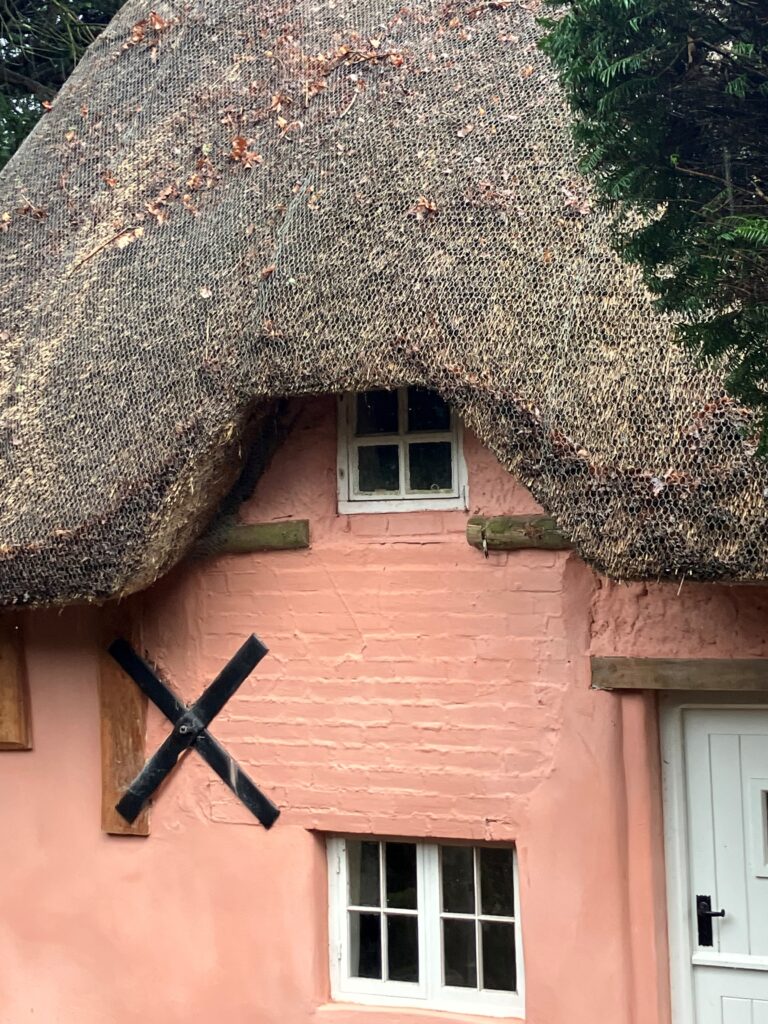 Close-up of pink cottage with steep thatched roof, small upper window, and black windmill-style wall feature beside white door
