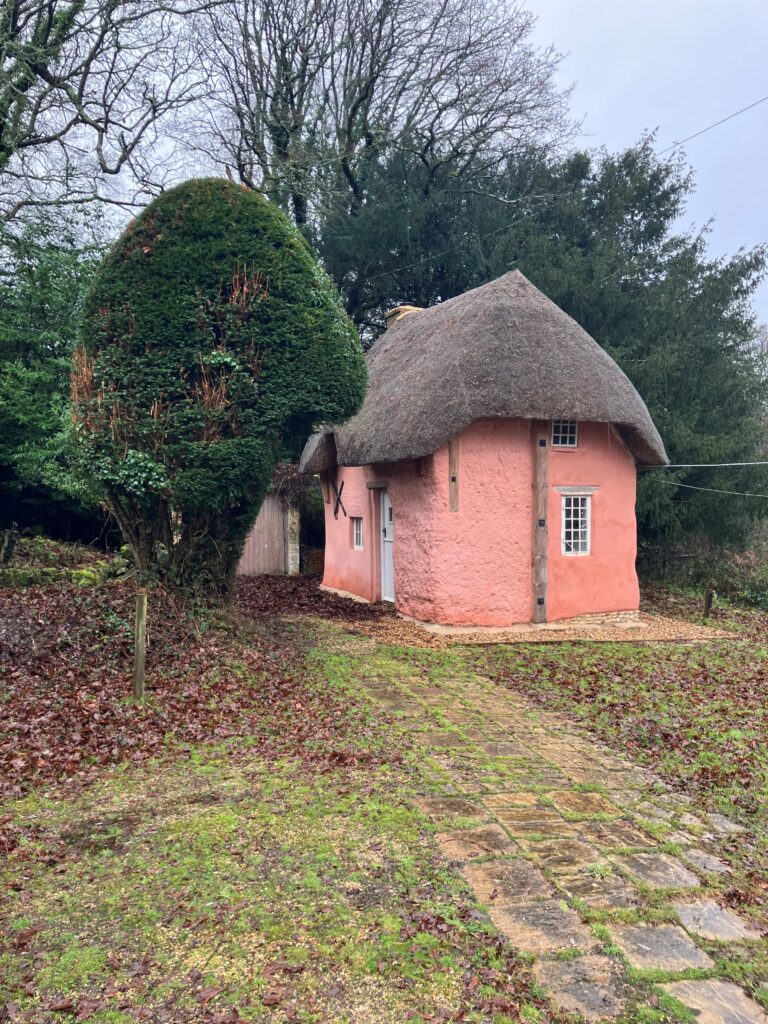 Restored pink thatched-roof cottage with white-framed windows, surrounded by trees and a stone path in a garden