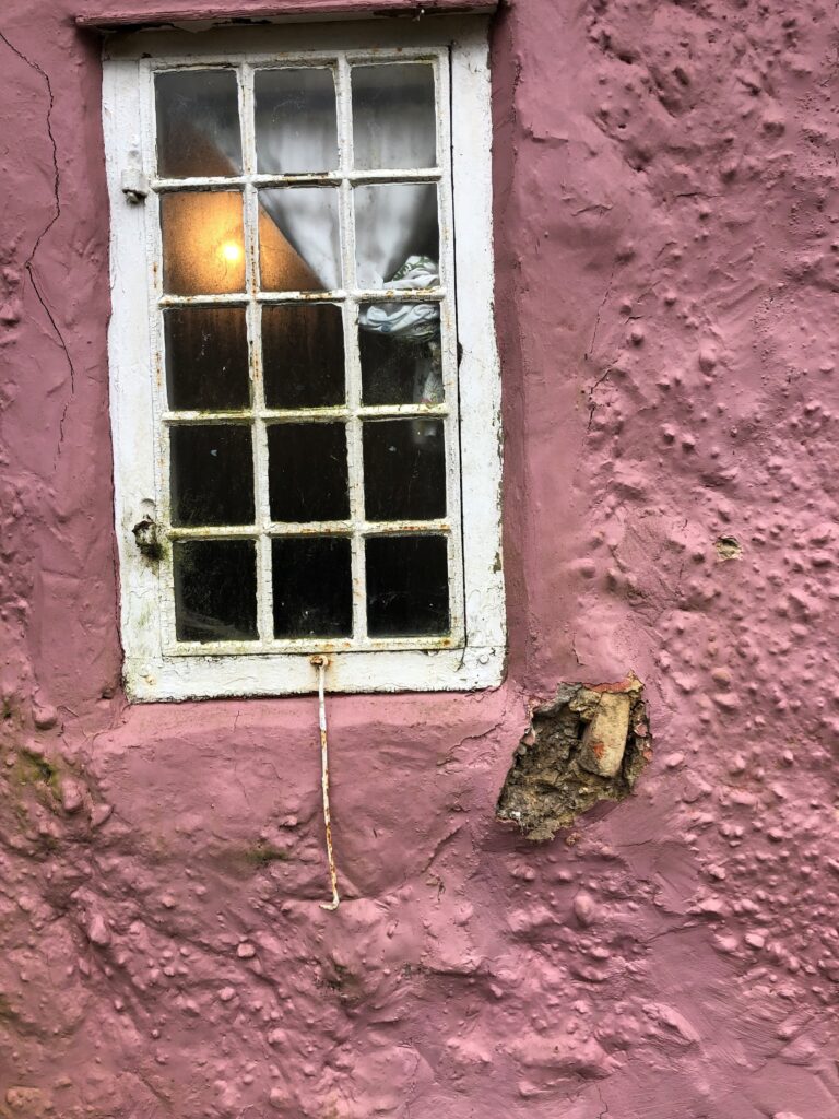 Weathered white-paned window in cracked pink stucco wall, with torn curtain, light glowing inside, and crumbling plaster below