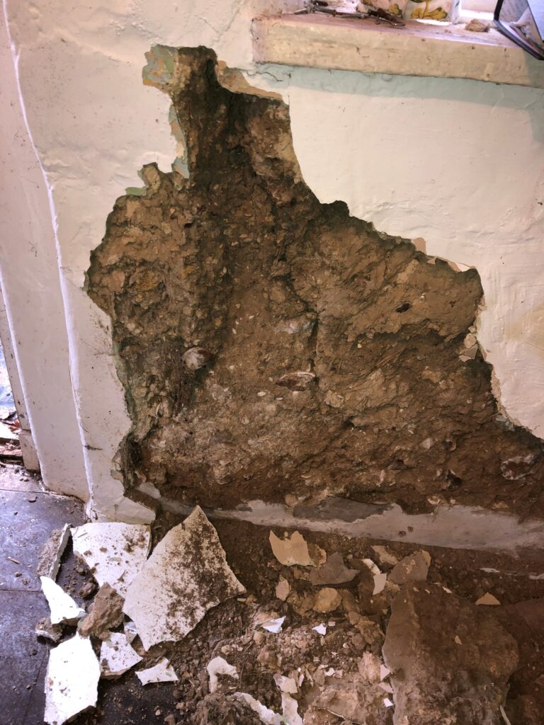 Crumbling interior wall with plaster missing, exposing rough stone, and rubble scattered on the floor beneath a windowsill