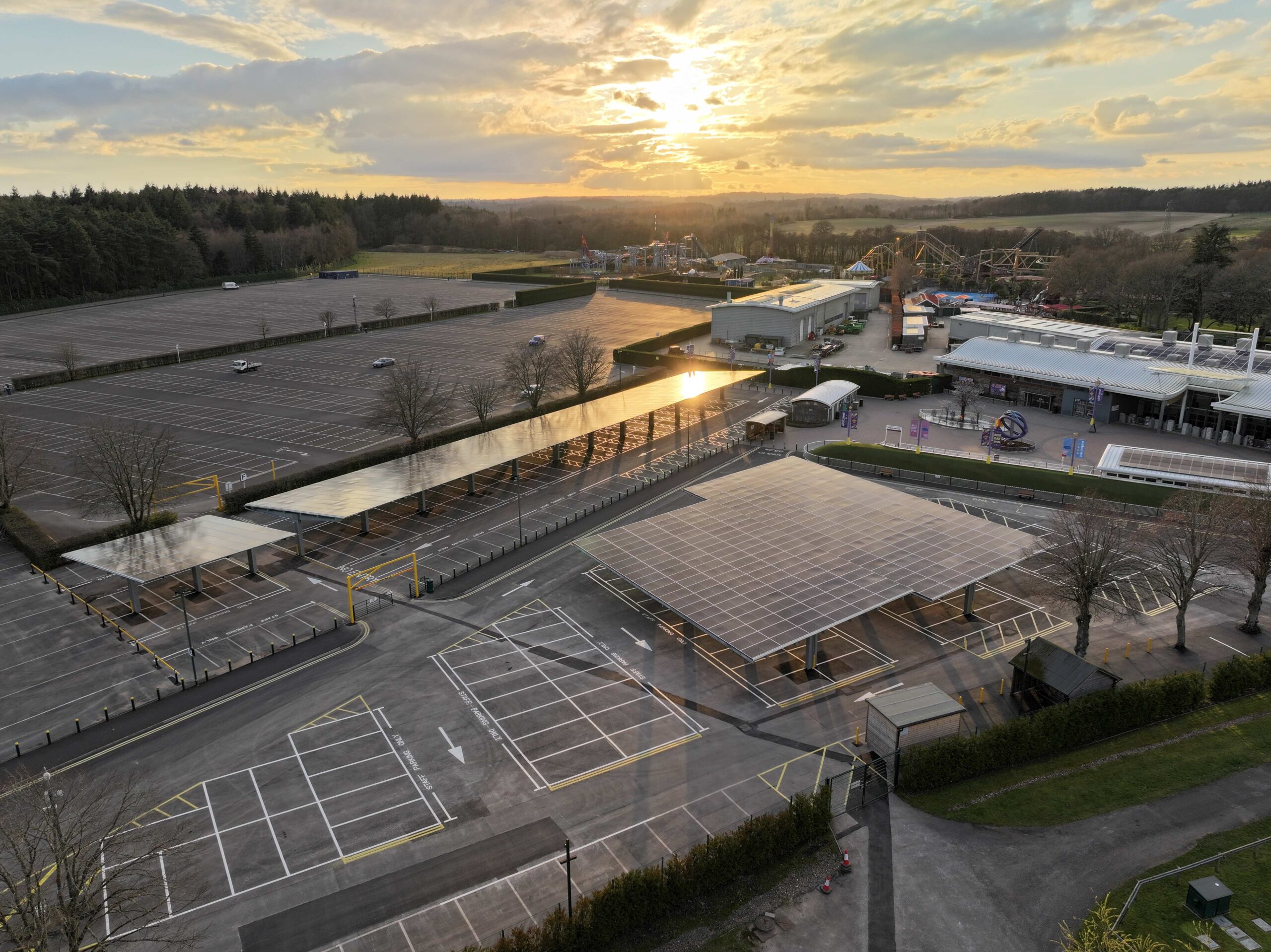 Solar panel carport canopies over Paultons Park parking spaces at sunset, with the park entrance building and rides in the distance