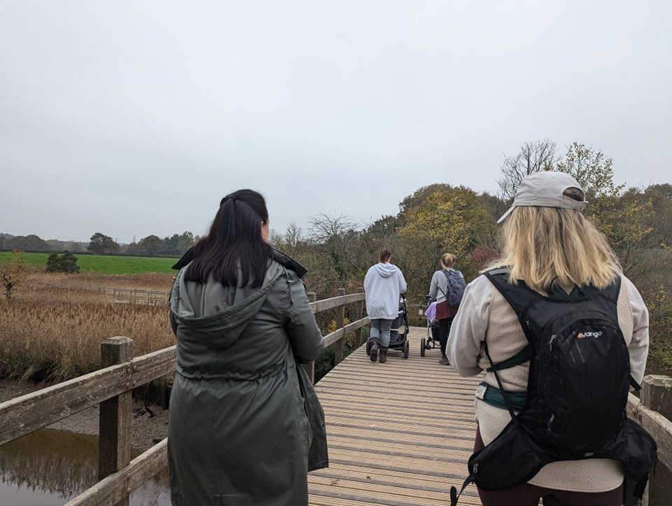 Group of people walking on a wooden boardwalk through wetlands, with trees and fields under an overcast sky