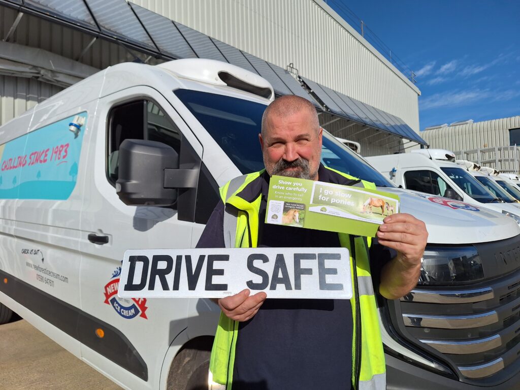 A man stood in front of a white van with a New Forest Ice Cream logo on the side. He is wearing hi vis and holding a numberplate reading Drive Safe as well as New Forest animal accident stickers and cards.