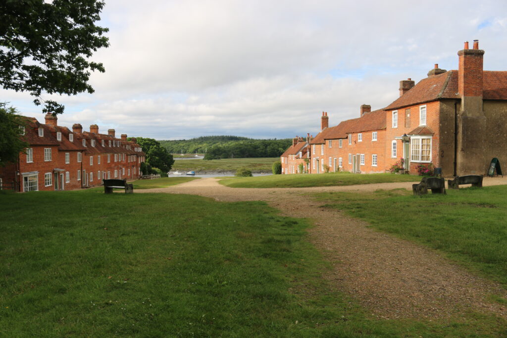 Grassy path between red-brick buildings at Buckler’s Hard, leading down to a river with small boats and wooded shore beyond
