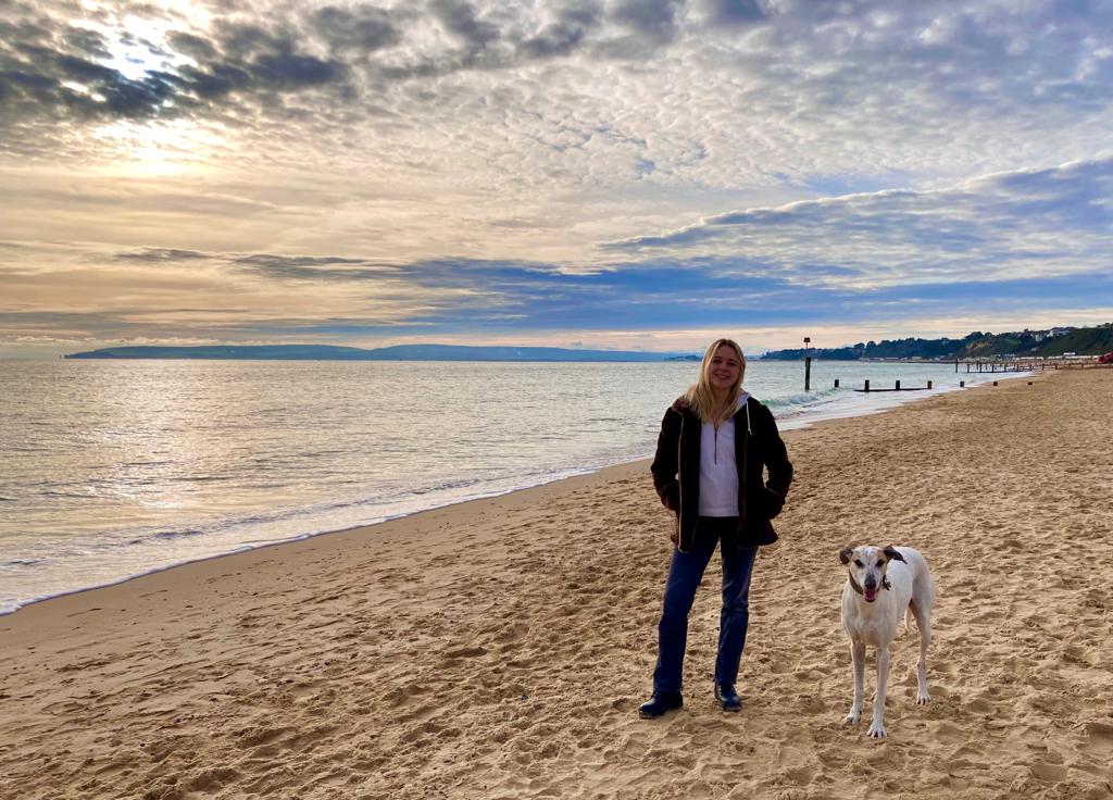 Person standing on sandy beach beside a white dog, calm sea and cloudy sunrise sky in background