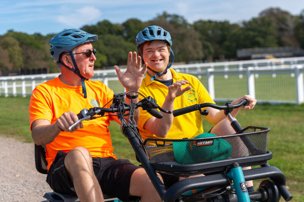 Two men on a side by side specialist bike. One is wearing a branded PEDALL New Forest Inclusive Cycling top