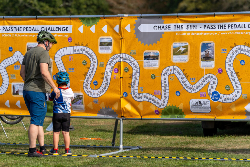 A father and son wearing cycle helmets stood in front of a large orange board depicting a winding path and distance markers. The board is covered in stickers showing how far people have cycled as part of a challenge.