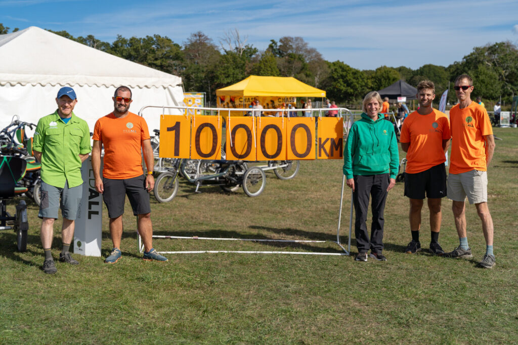 A group of people stood by a large distance counter showing 10,000. They are at an outdoor cycling event on a sunny day.